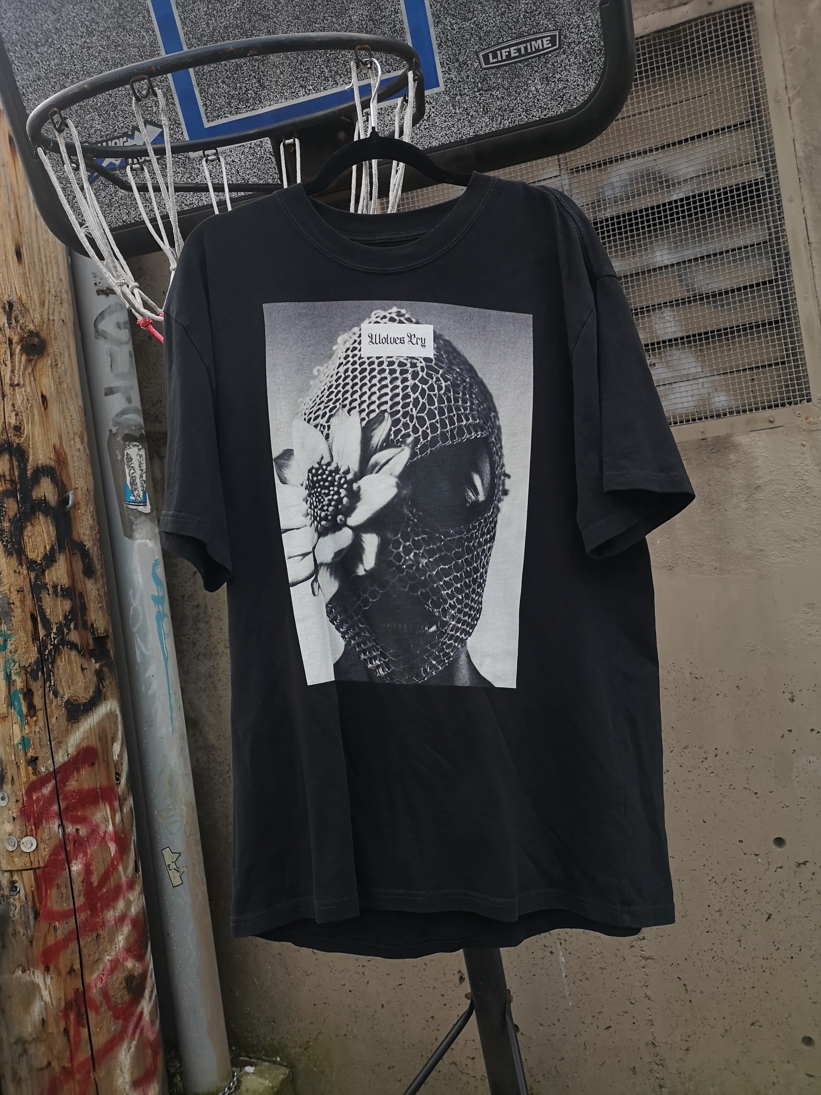 Wolves Cry Oversized Balaclava Tee in vintage black displayed hanging, with its graphic design prominently featured against an urban background.