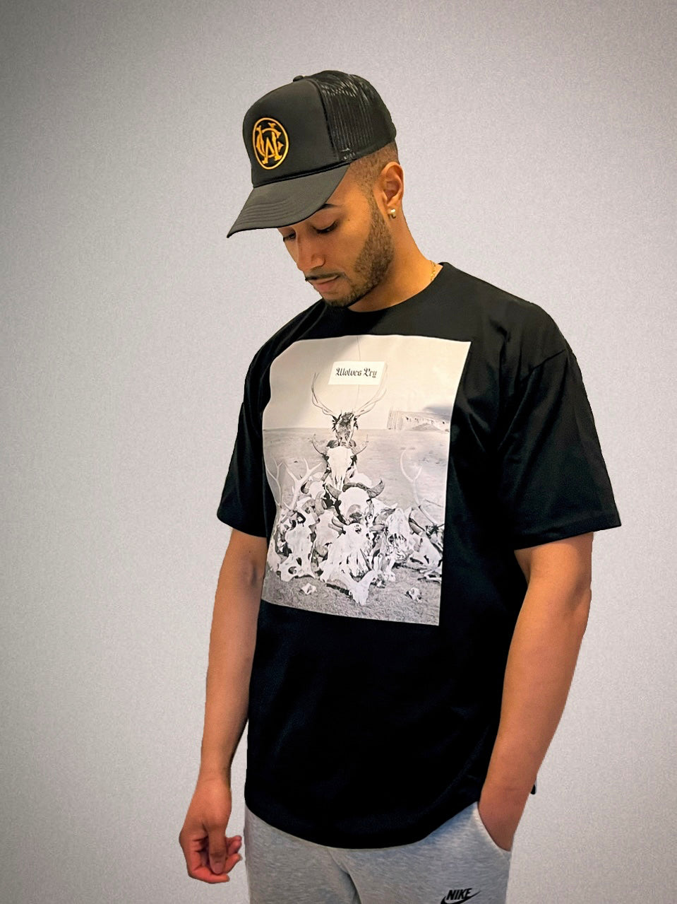 Male model wearing Wolves Cry Death Valley Tee in black, styled with a black trucker hat. Modern streetwear look with a bold monochrome graphic.